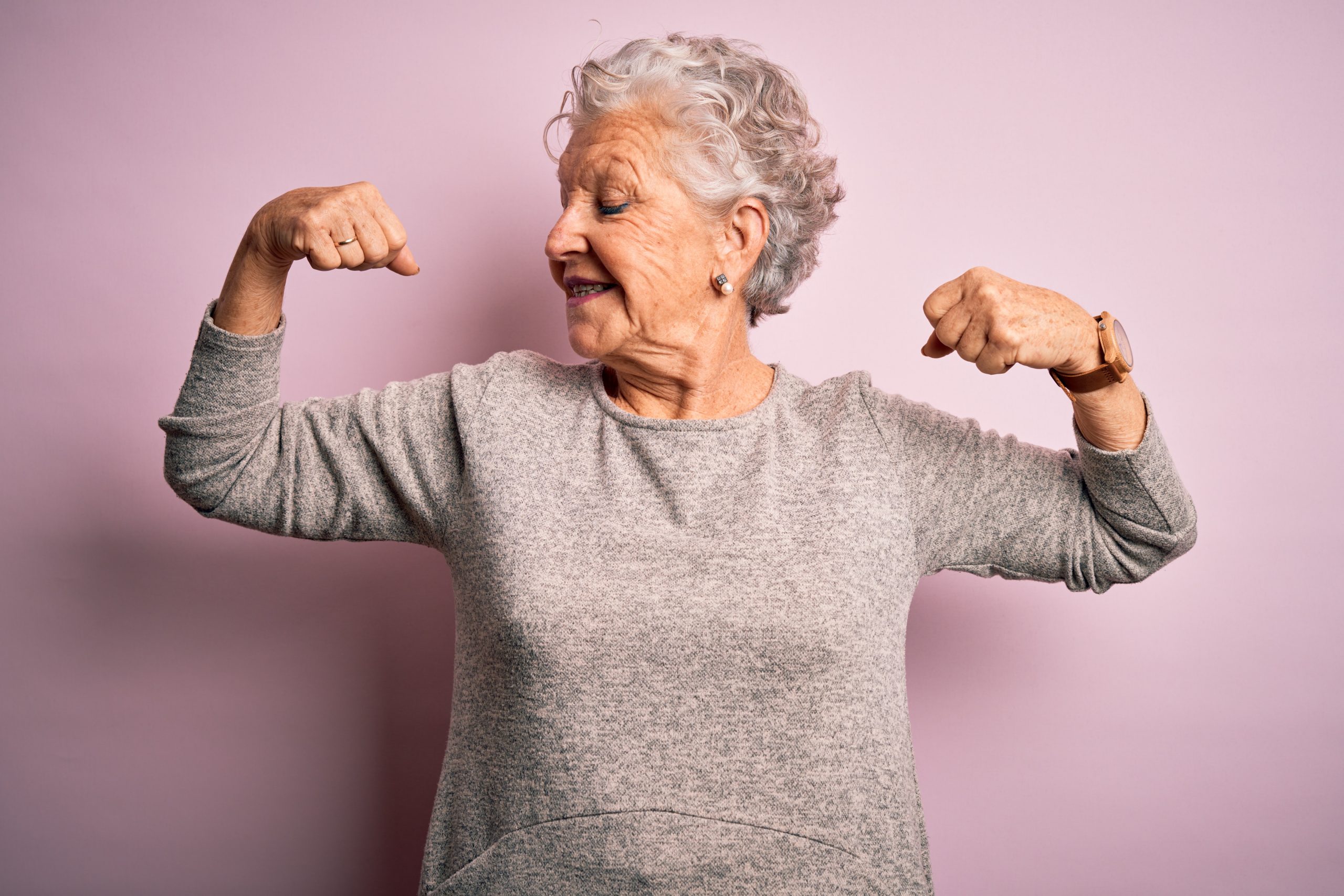 Senior beautiful woman wearing casual t-shirt standing over isolated pink background showing arms muscles smiling proud. Fitness concept.